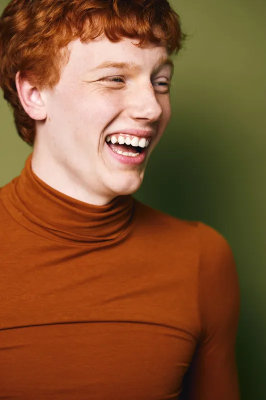 Young man with red hair and freckles laughing with teeth visible wearing brown turtleneck sweater on green background. Happy expression, casual portrait, smiling and youthful person.