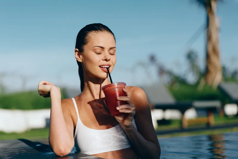 Young woman in a white swimsuit relaxes in a bright poolside scene, sipping a red smoothie through a straw and enjoying a carefree summer moment by the water.