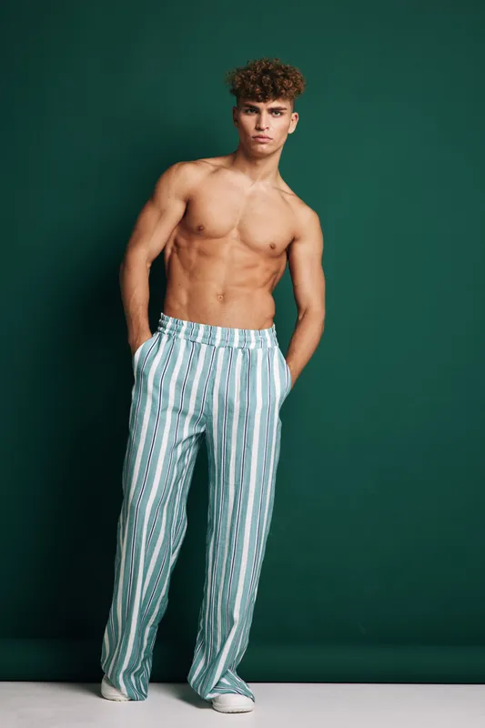 Young man posing shirtless wearing striped pants and white sneakers against green wall. Fashion shot highlights casual clothing, toned body, confident pose and studio lighting for model look.