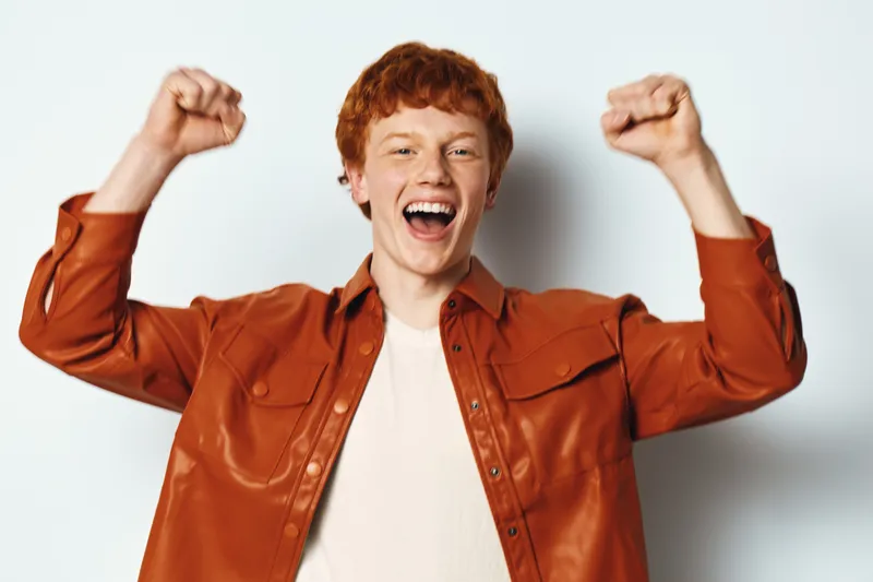 Young man with red hair in orange shirt raising fists and celebrating with joyful expression on white background for happiness and success emotions.