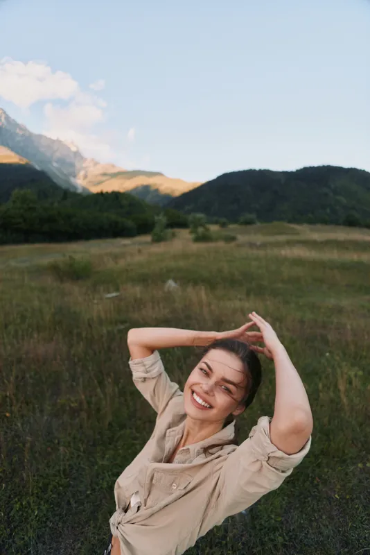 A smiling woman enjoys outdoor lifestyle in a field with distant mountains, capturing a carefree moment in nature in a sunlit meadow, wearing casual attire.