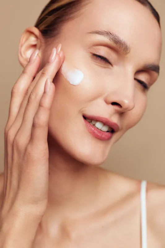 A young woman applies moisturizing cream to her cheek with closed eyes in a close-up portrait. The soft beige background highlights skincare and beauty care concept.