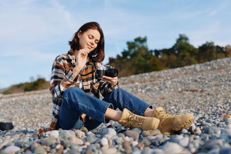 Woman photographer sits on a rocky beach, wearing a plaid shirt and jeans, holding a camera and smiling as she reviews shots by the shoreline under a bright sky, enjoying a calm outdoor moment