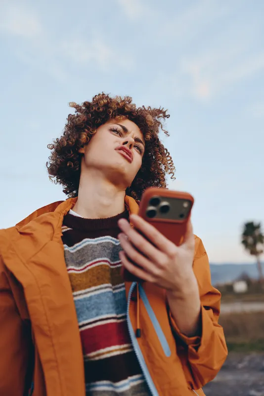 Woman with curly hair holding a smartphone for a selfie outdoors in bright blue sky, wearing an orange jacket and striped sweater, Lifestyle moment in a calm countryside setting