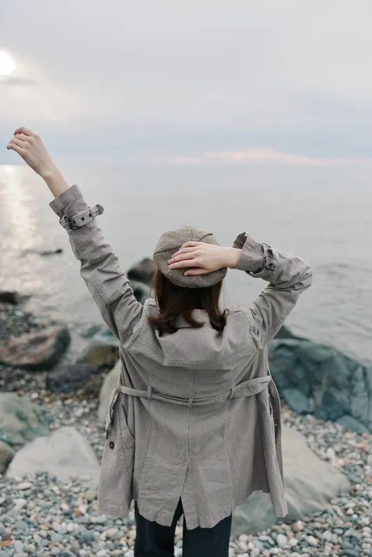 Woman wearing coat and hat stands on rocky shore facing sea with raised arm. Overcast sky creates calm mood. Concept of freedom, nature, and peaceful outdoor moment.