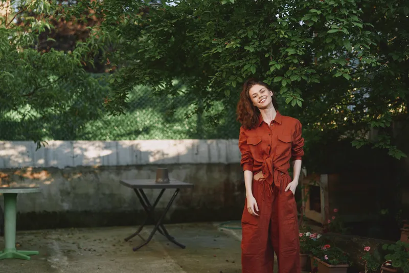 A smiling woman in a red jumpsuit stands in a sunlit garden, relaxed pose and casual confidence. Outdoor lifestyle portrait expressing warmth, ease, and everyday fashion