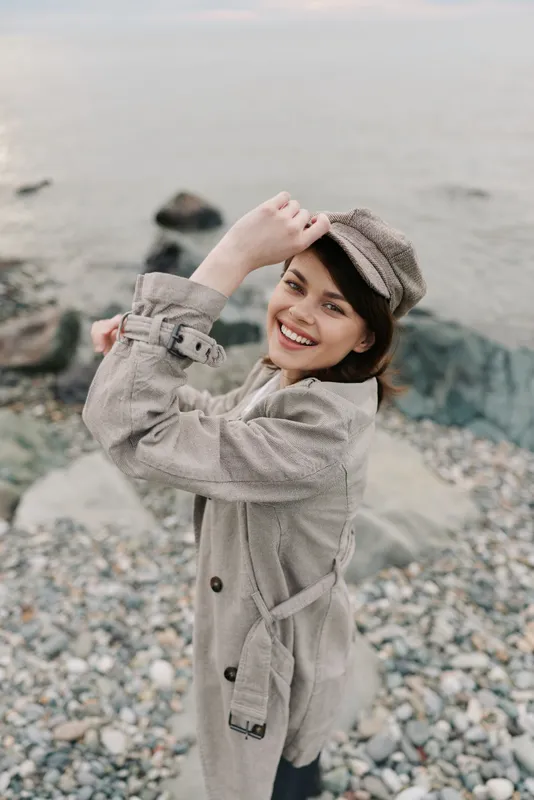 Smiling young woman in trench coat and hat poses happily on rocky beach. Natural light highlights her joyful expression and casual autumn style by the sea. Concept of outdoor fashion.