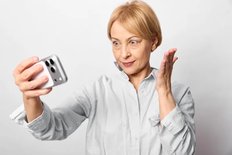 Woman senior takes selfie with smartphone, technology and communication concept, smiling portrait in white studio lighting, relaxed pose, peace gesture