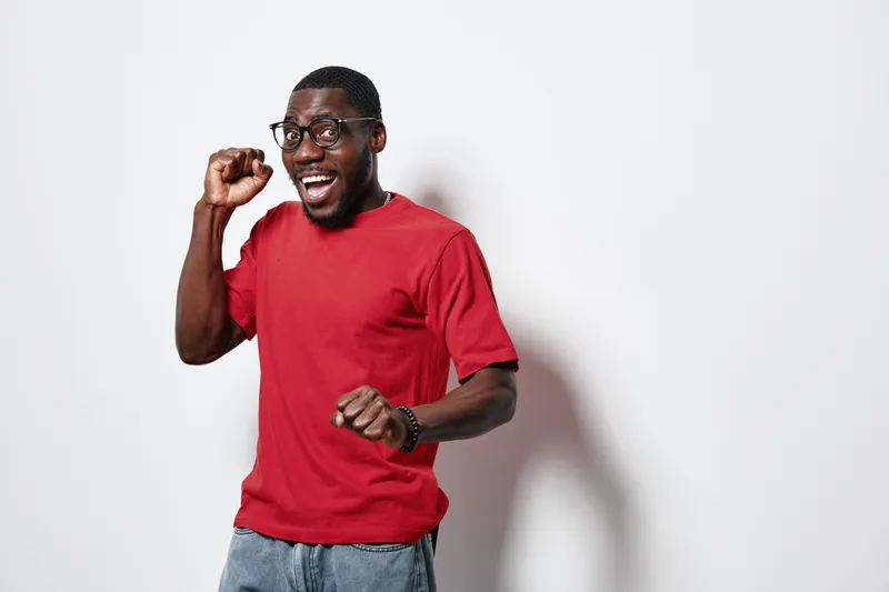 African American man with dark skin wearing glasses and red t shirt celebrating success with raised fist and joyful expression on white background in studio.