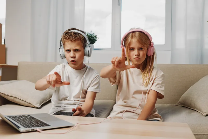 Two kids with headphones sitting on sofa making finger gestures at camera, using laptop in cozy living room with natural light and neutral decor.