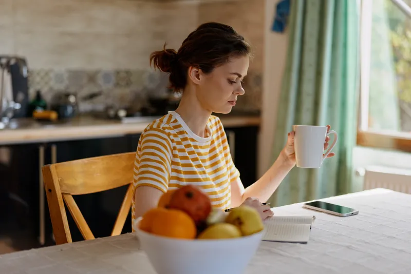 A woman sits at a bright kitchen table, sipping from a mug and glancing at a phone, while a bowl of fruit rests nearby in soft morning light. Her striped shirt adds warmth to the scene and the open
