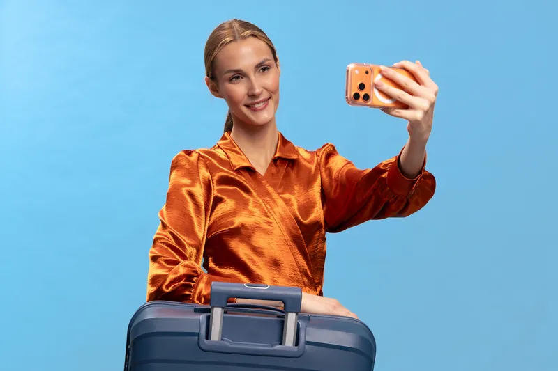 Young woman in orange blouse takes selfie with smartphone while holding blue suitcase on blue background. Concept for travel, vacation preparation, and modern lifestyle.