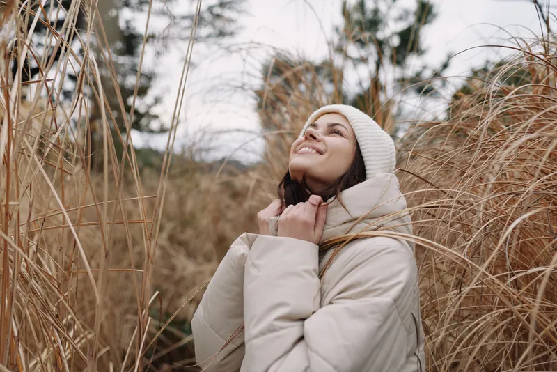 Woman in a warm coat and scarf standing among tall grass, looking up with a serene smile in a chilly outdoor landscape scene