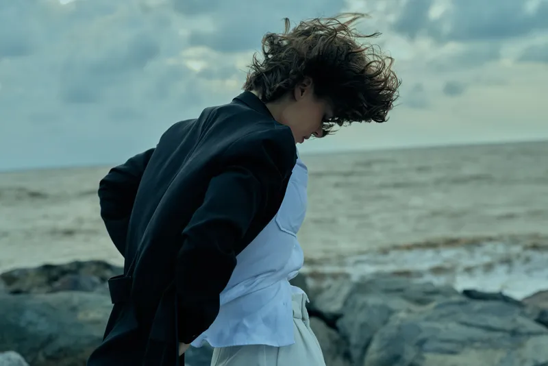 Windblown hair and a dark jacket paired with a light shirt beside the seashore, rocks and ocean under a moody sky, an outdoor fashion moment with natural movement