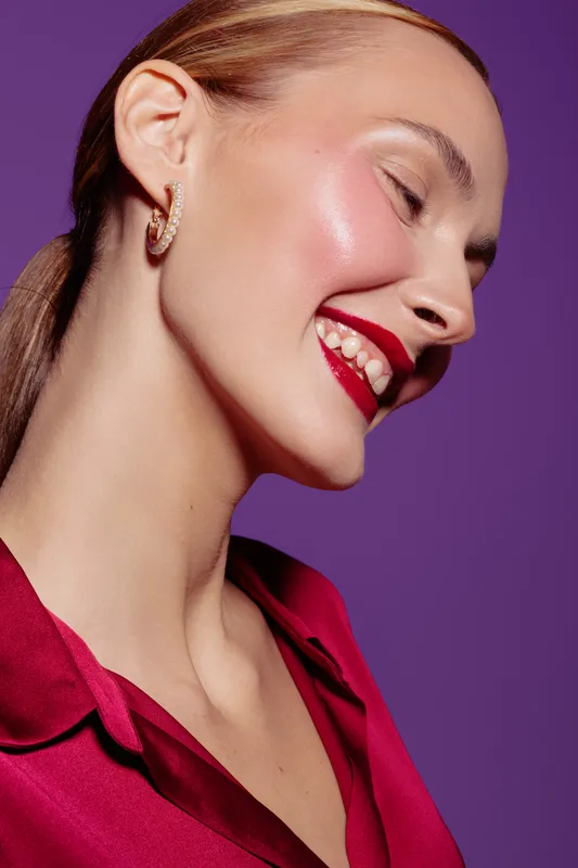 Smiling woman with red lipstick and shiny skin poses in red blouse against purple background. Concept of beauty, makeup, fashion, and confident female portrait in studio lighting.