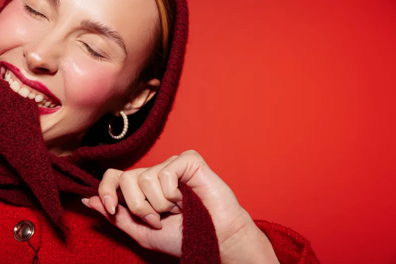 Smiling woman in red sweater with hood closes eyes and holds collar, enjoying cozy moment. Bright red background creates warm and joyful atmosphere of comfort and happiness.