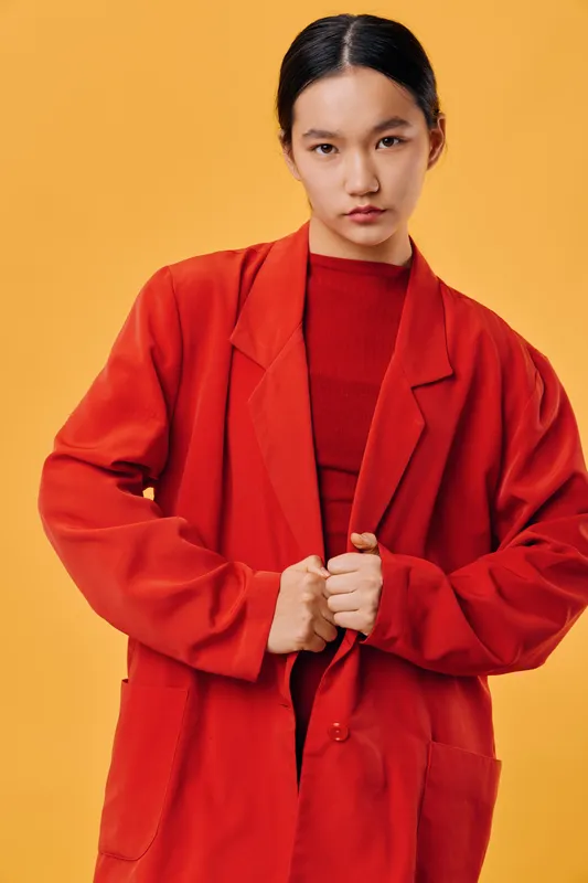 Young woman in oversized red coat and matching sweater posing confidently against orange background. Fashion portrait with minimalist style and intense look, model wearing casual outerwear indoors.