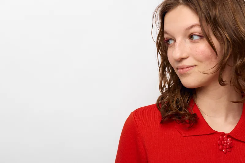 Young woman with curly brown hair wearing red sweater looks sideways with slight smile on white background. Concept of casual style, natural beauty, and positive mood.