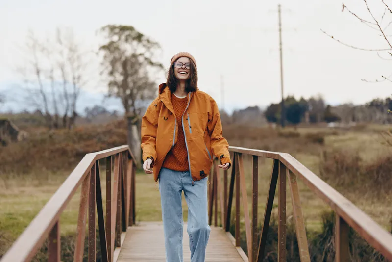Smiling woman walking outdoors on a wooden bridge in autumn, casual lifestyle portrait in nature park, jeans and orange jacket, soft morning sunlight over trees and path, calm wellbeing mood