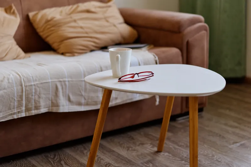 Cozy living room scene featuring a white round coffee table with a ceramic mug and red glasses resting on its surface, a soft beige sofa in the background.
