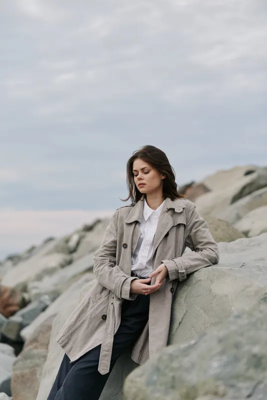 Young woman in a textured coat leans against rocks outdoors, looking down thoughtfully. Overcast sky and natural rocky background create a calm, contemplative mood in the image.
