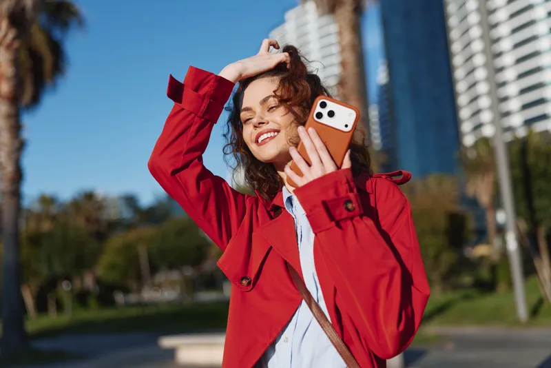 Cheerful woman outdoors, selfie with a red jacket, phone in hand, sunny urban scene, smiling expression, casual fashion moment in daylight