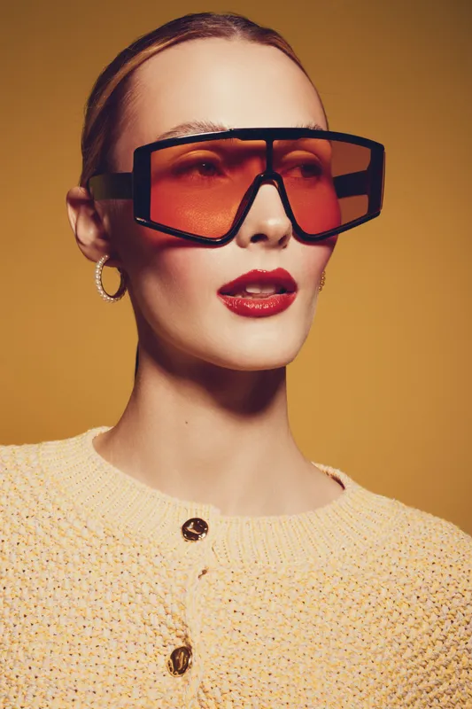 Stylish woman with oversized orange sunglasses and red lipstick poses against warm brown background. Fashion portrait with trendy eyewear and elegant knitted sweater. Modern beauty concept.