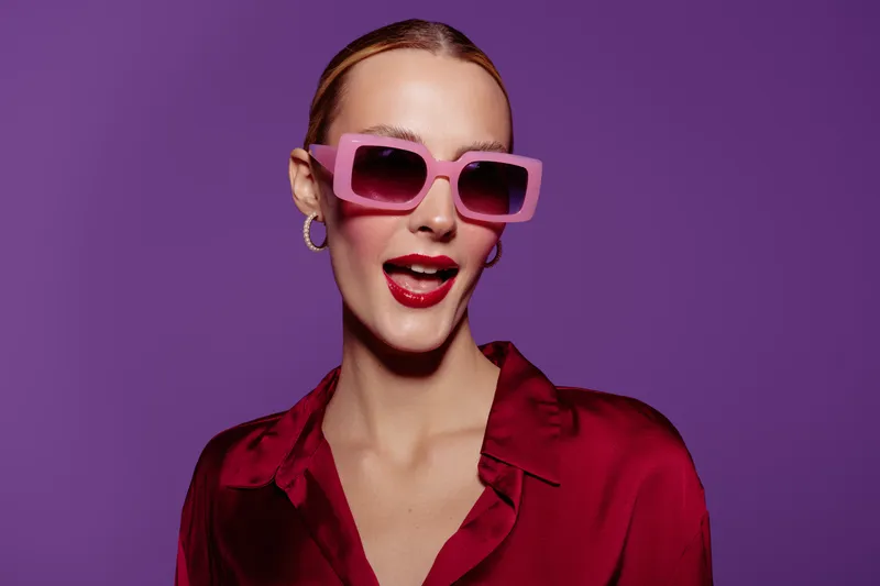 Stylish woman with slicked back hair wears oversized pink sunglasses and red lipstick. She poses confidently against a purple background in a red satin blouse. Fashion and beauty concept.