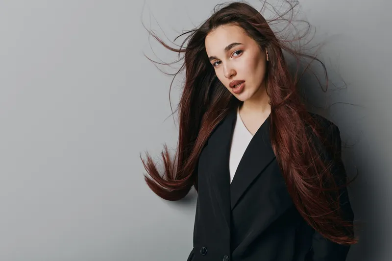 Woman with long hair wearing black blazer posing against gray background, looking at camera with confident expression and natural makeup in studio portrait.