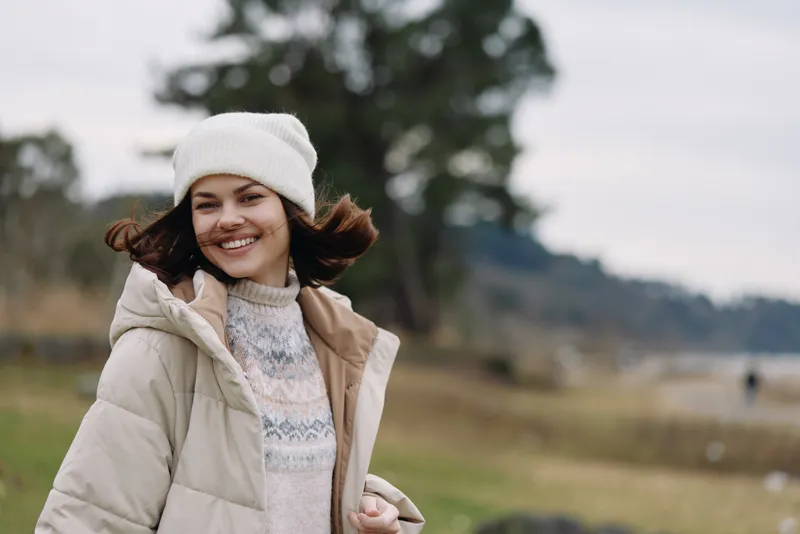 Woman in a winter coat and beanie smiles outdoors in a breezy park, soft natural light, cozy fashion, chilly weather, casual pose, calm landscape behind