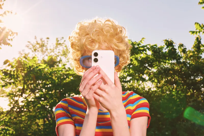 Woman with curly blonde hair wears a rainbow striped top and blue sunglasses, taking a selfie outdoors under bright sun, retro film color vibe, casual lifestyle moment.