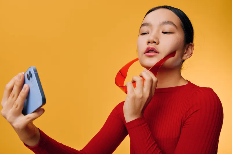 Young woman taking selfie with smartphone on orange background wearing red sweater holding face mask in hand, casual portrait and modern lifestyle with health awareness concept.