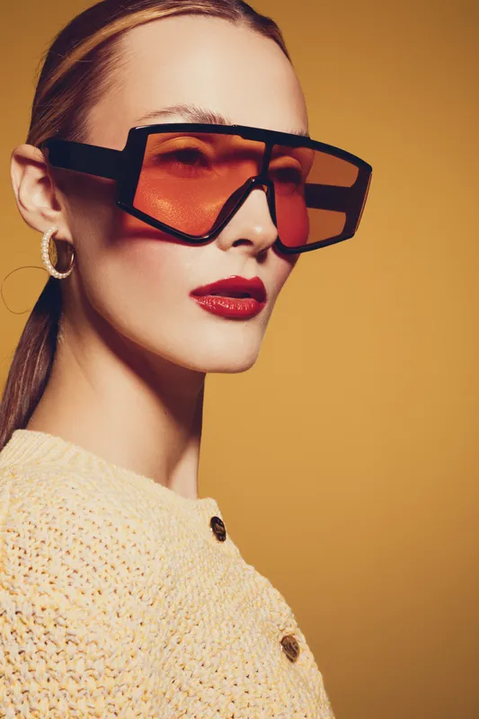 Stylish woman with oversized orange sunglasses and red lipstick poses against yellow background. Fashion portrait with modern accessories and confident expression.