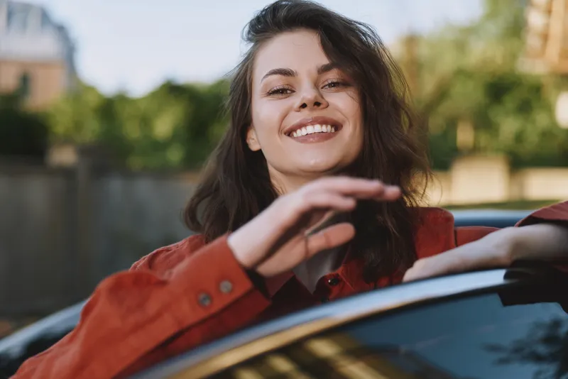 Happy woman leaning out of a car window on a sunny day, showcasing casual style, carefree lifestyle, natural smile, urban outdoor setting, candid portrait, joyful mood