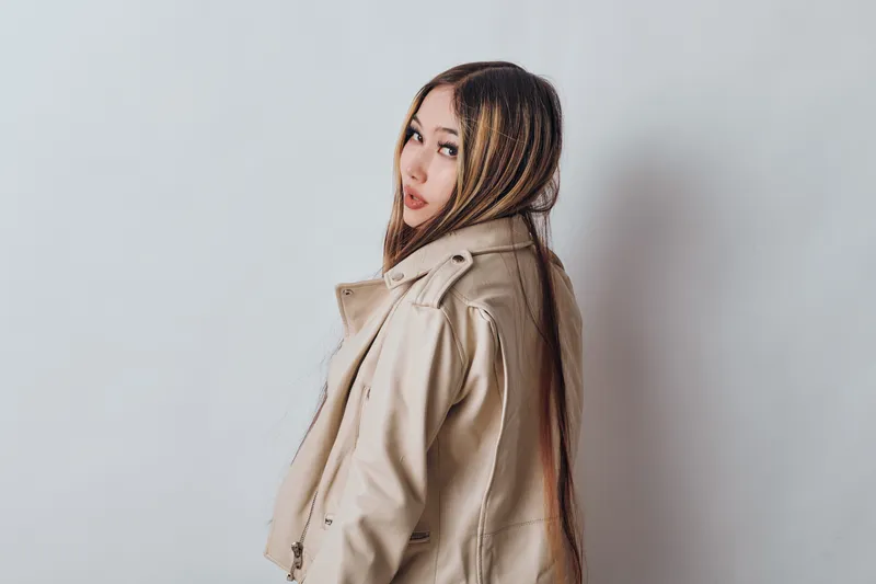 Young woman with braided hair wearing a beige jacket, posing sideways on a plain light background. Fashion portrait with casual style and long hairstyle in studio.