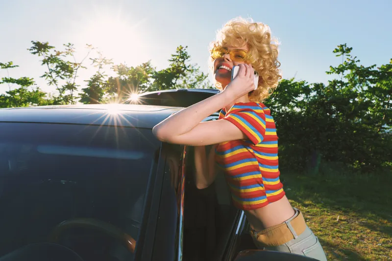 A stylish woman in a striped crop top and blonde wig leans on a dark car in bright sunlight, talking on a mobile phone. A playful retro vibe meets a carefree summer lifestyle.