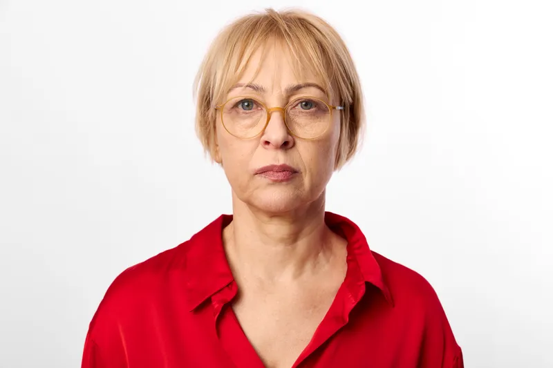 Senior woman, red shirt, glasses, studio portrait, head and shoulders, neutral expression, looking at camera, mature face, calm mood, white background, soft even light