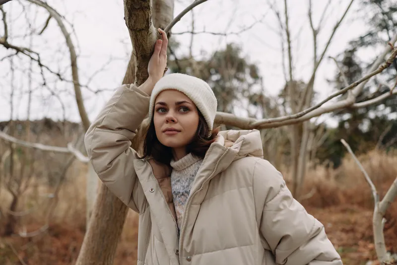 Woman in a beige winter jacket and knit beanie leans beside a tree in a bare woodland, calm pose, soft natural light, moody outdoor portrait, with textured fabric and subtle shadows. Soft light.