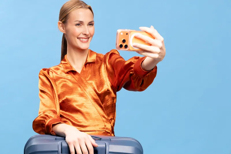 Young woman in orange blouse takes selfie with smartphone, sitting on suitcase. Concept for travel, vacation, lifestyle, and social media communication on blue background.