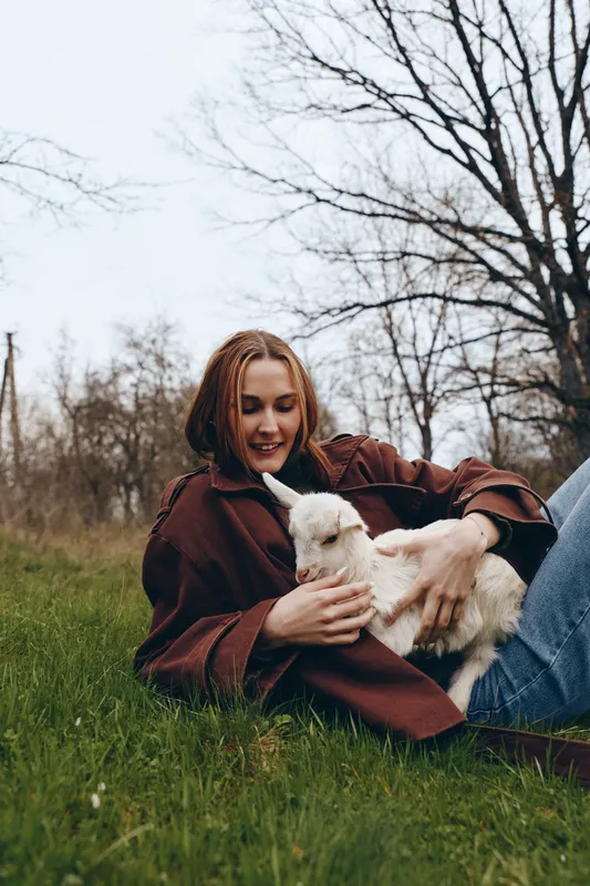 Joyful woman in a brown coat lies on grass outdoors, gently cuddling a white goat kid, capturing a warm lifestyle moment in a park setting with natural light and relaxed mood.