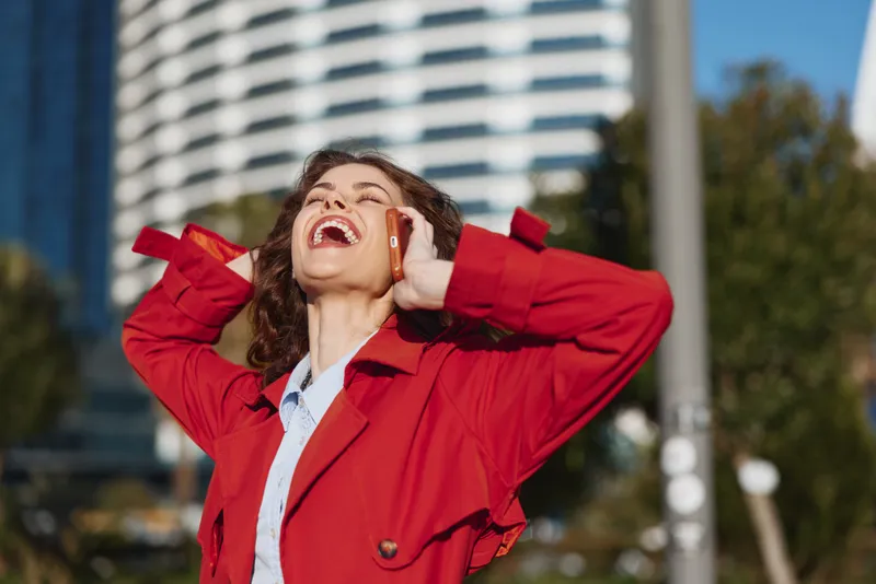 Young woman with curly hair wearing a red coat laughs joyfully while talking on the phone outdoors in an urban setting on a sunny day.