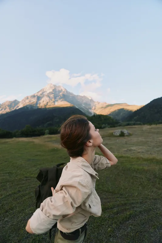 Young woman with a backpack gazes at distant mountains, enjoying a moment of awe during a hike in an expansive alpine meadow, sunlight warming the scene.