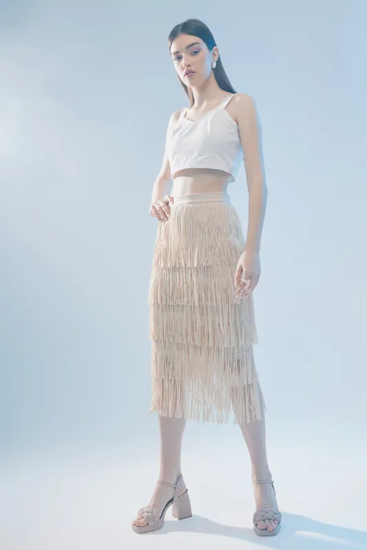 Young woman in a white crop top and beige fringed skirt poses confidently in a studio with soft lighting. Concept of woman beauty fashion and modern style.