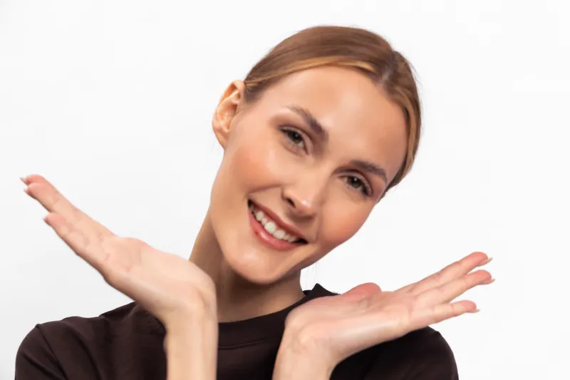 Smiling young woman with blonde hair and natural makeup poses with hands near face on white background. Concept of happiness, beauty, youth, and positive emotion in studio portrait.