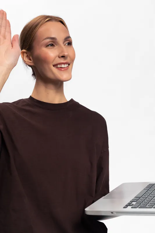 Smiling woman with light hair holds laptop and waves hand, isolated on white background. Concept for online communication, video call, remote work, and friendly interaction.