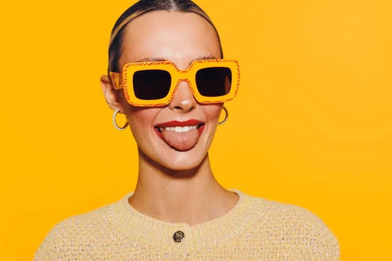 Young woman with orange sunglasses and hoop earrings sticks out tongue on bright yellow background. Casual style, playful mood, and summer fashion concept with vibrant colors and fun expression.