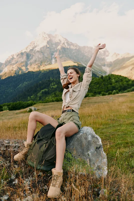 A young woman sits on a rock in a mountain meadow, arms raised in triumph, a backpack beside her, capturing a candid moment of outdoor adventure and pure joy.