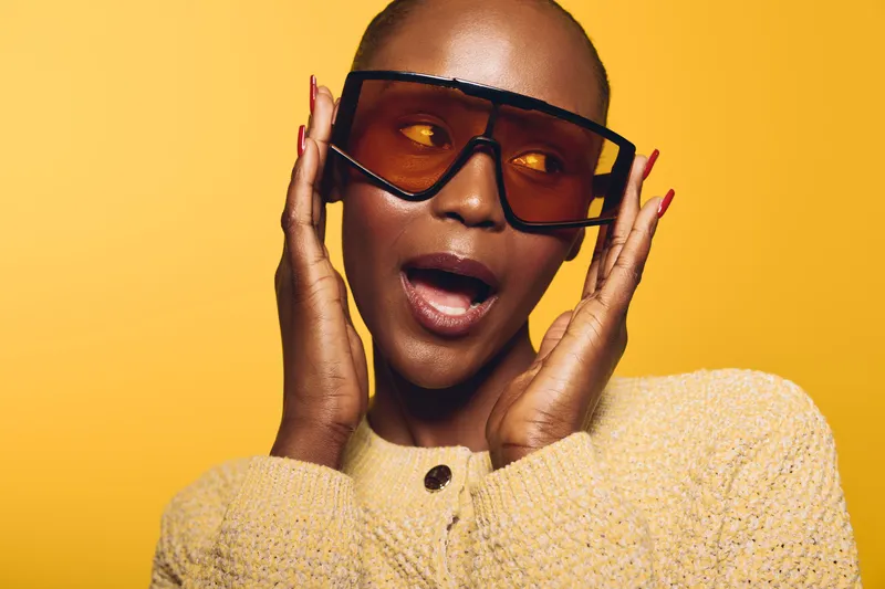 Portrait of a dark skinned African woman wearing oversized sunglasses and a beige knit sweater against a yellow background. Concept of fashion, style, and confident beauty.