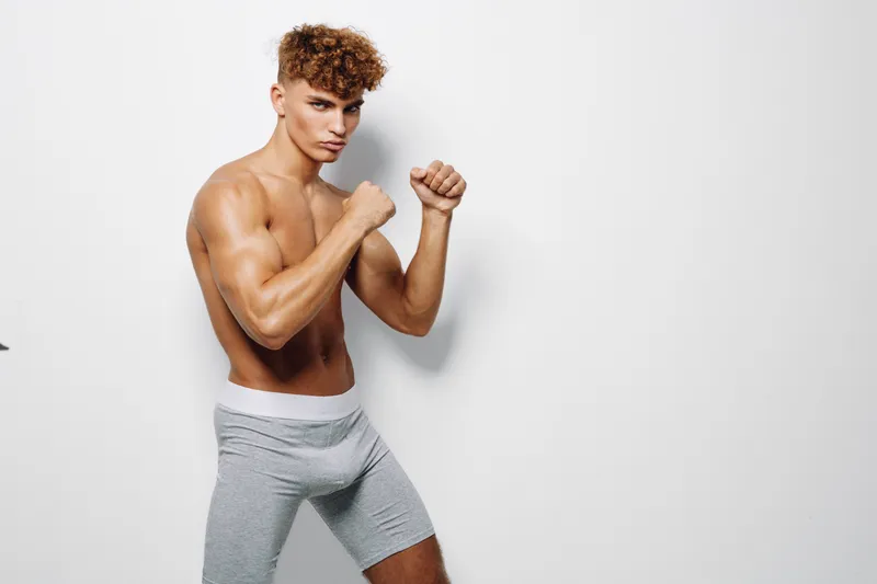 Young muscular man with curly hair posing shirtless in fighting stance wearing gray shorts against white background for fitness and boxing concept.