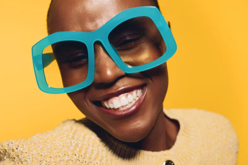 Close-up of happy African woman with dark skin wearing large blue glasses and yellow sweater smiling brightly against yellow background. Concept of joy, fashion, and positive emotions.
